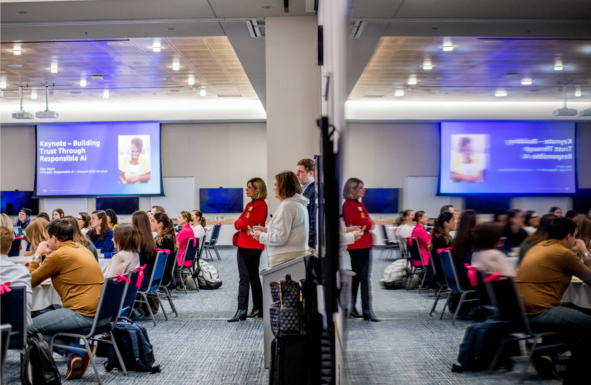 Conference room with mirrored reflection showing seated attendees and a woman in a red jacket.